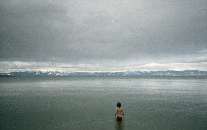 A person stands in a vast lake with snow-capped mountains in the distance under a cloudy sky.