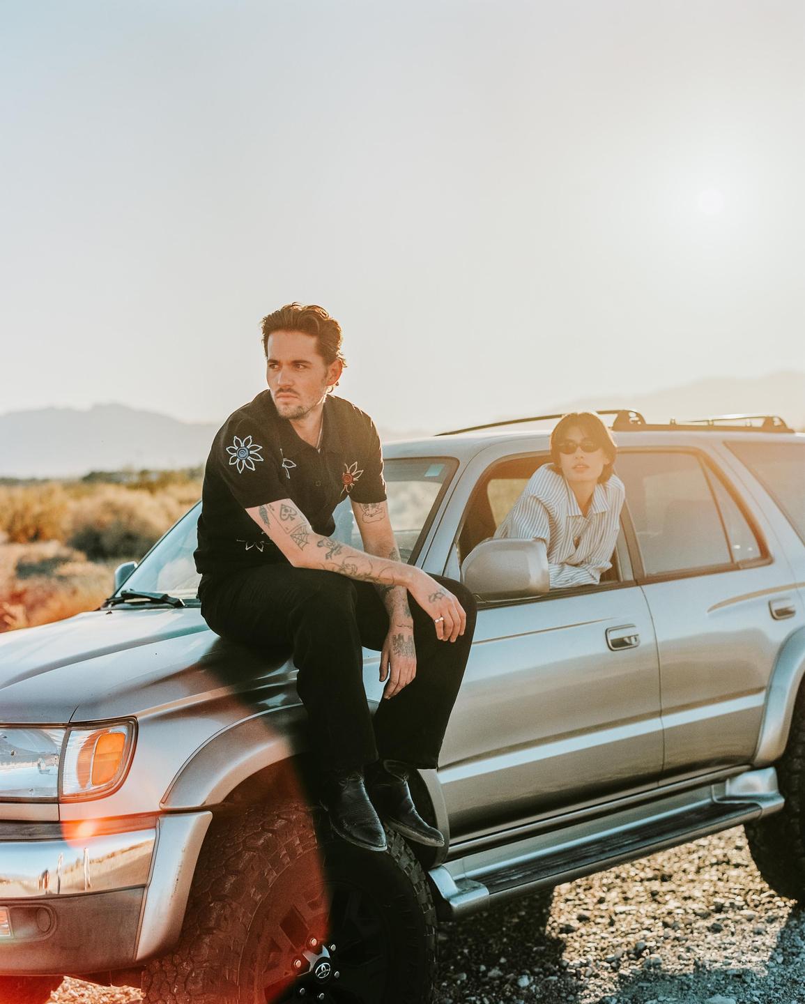 A man sits on the hood of a silver SUV while a woman looks out the passenger window at golden hour.