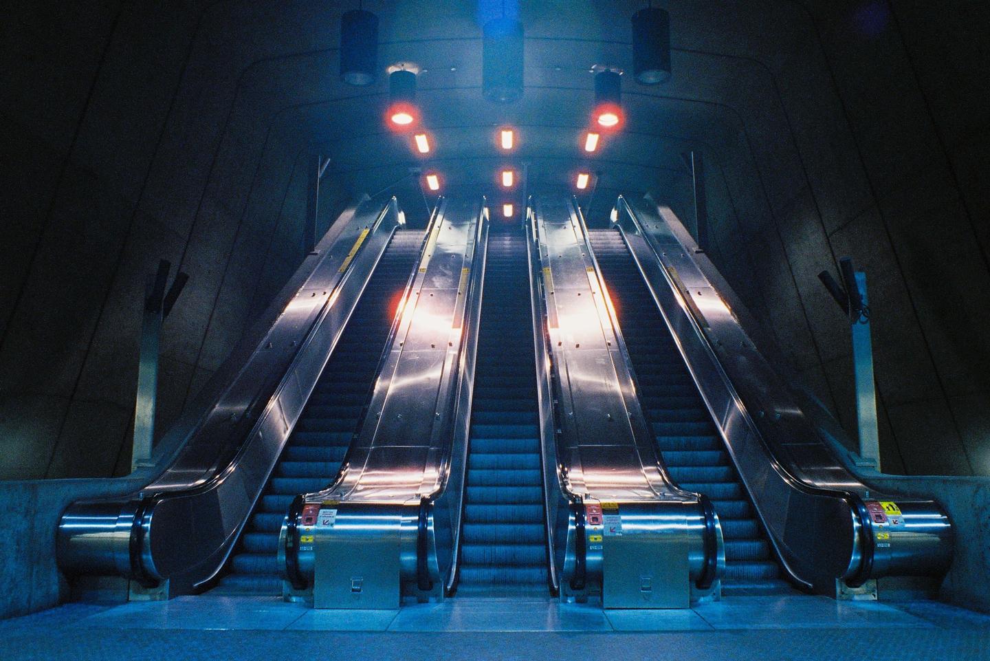 Two escalators ascend into a dark, cavernous space illuminated by red and blue lights.