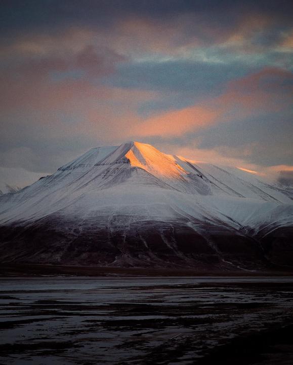 A snow-covered mountain peak is illuminated by golden light under a cloudy sky.