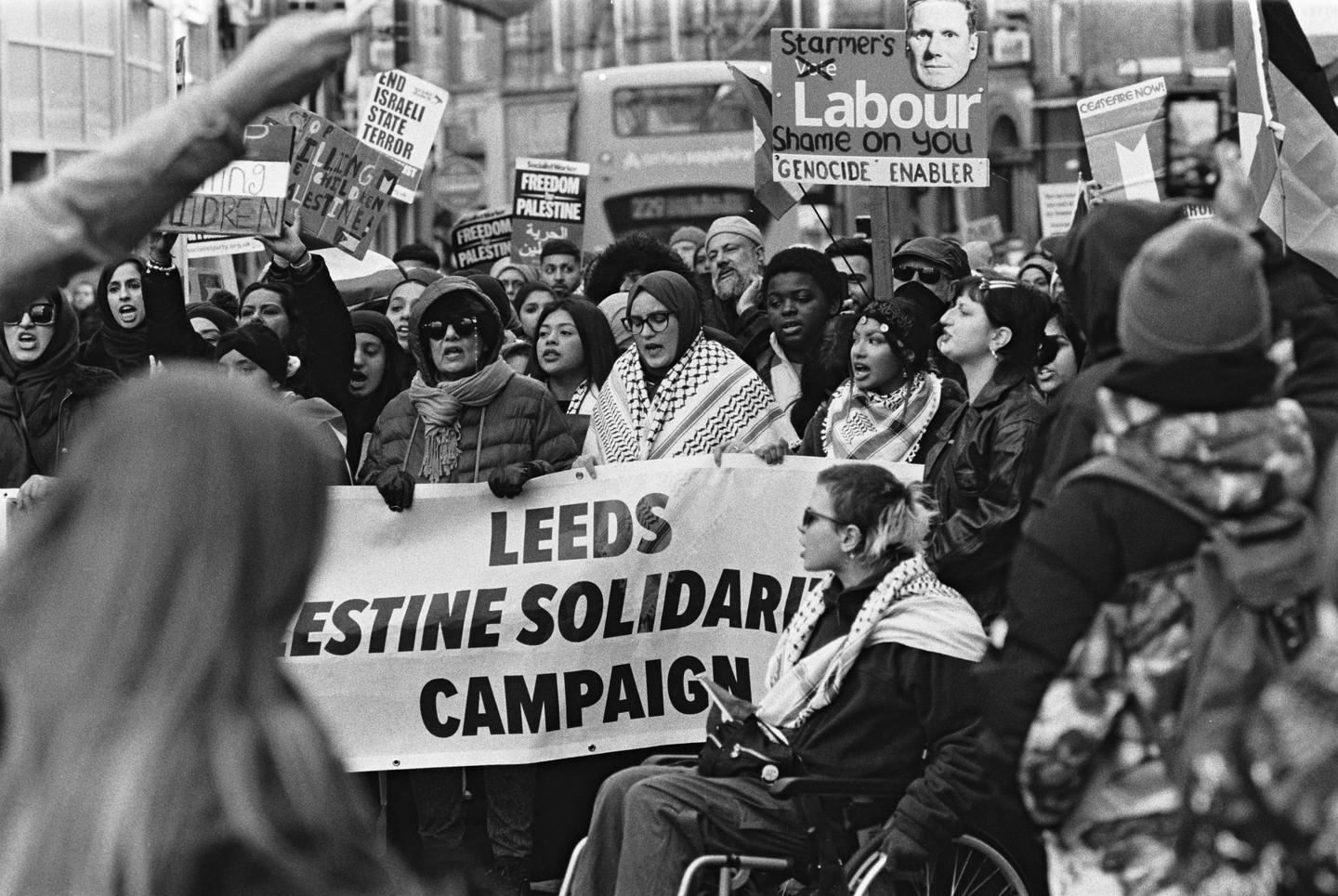 A crowd of people protest in a street, holding signs and a banner.