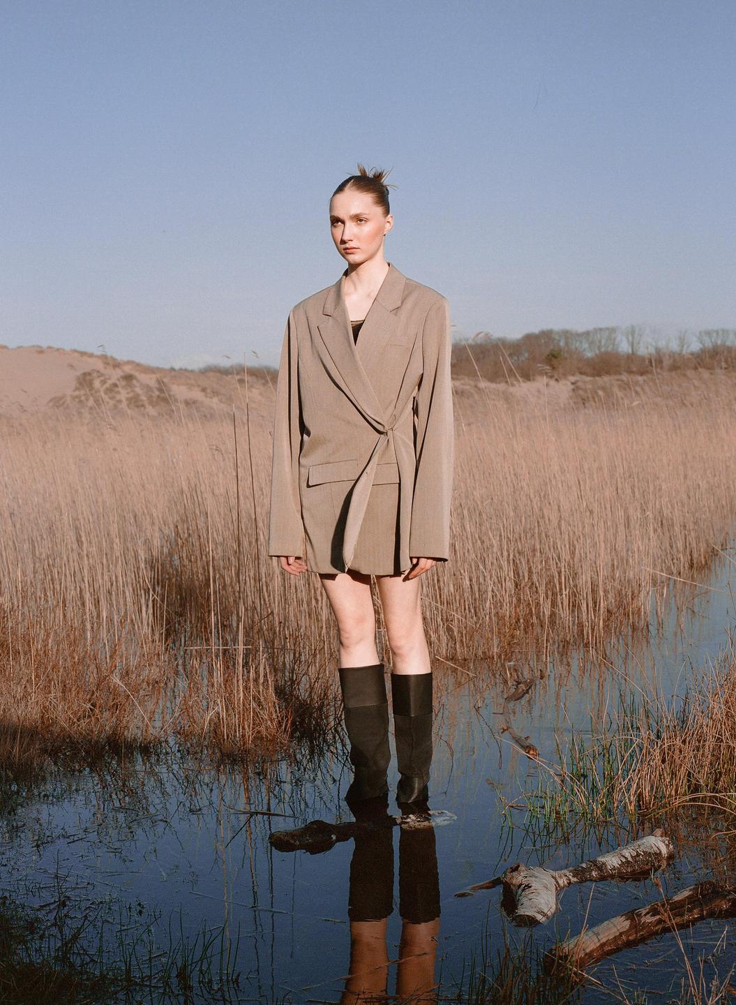 A woman in a blazer and tall boots stands in shallow water surrounded by tall dry grass.