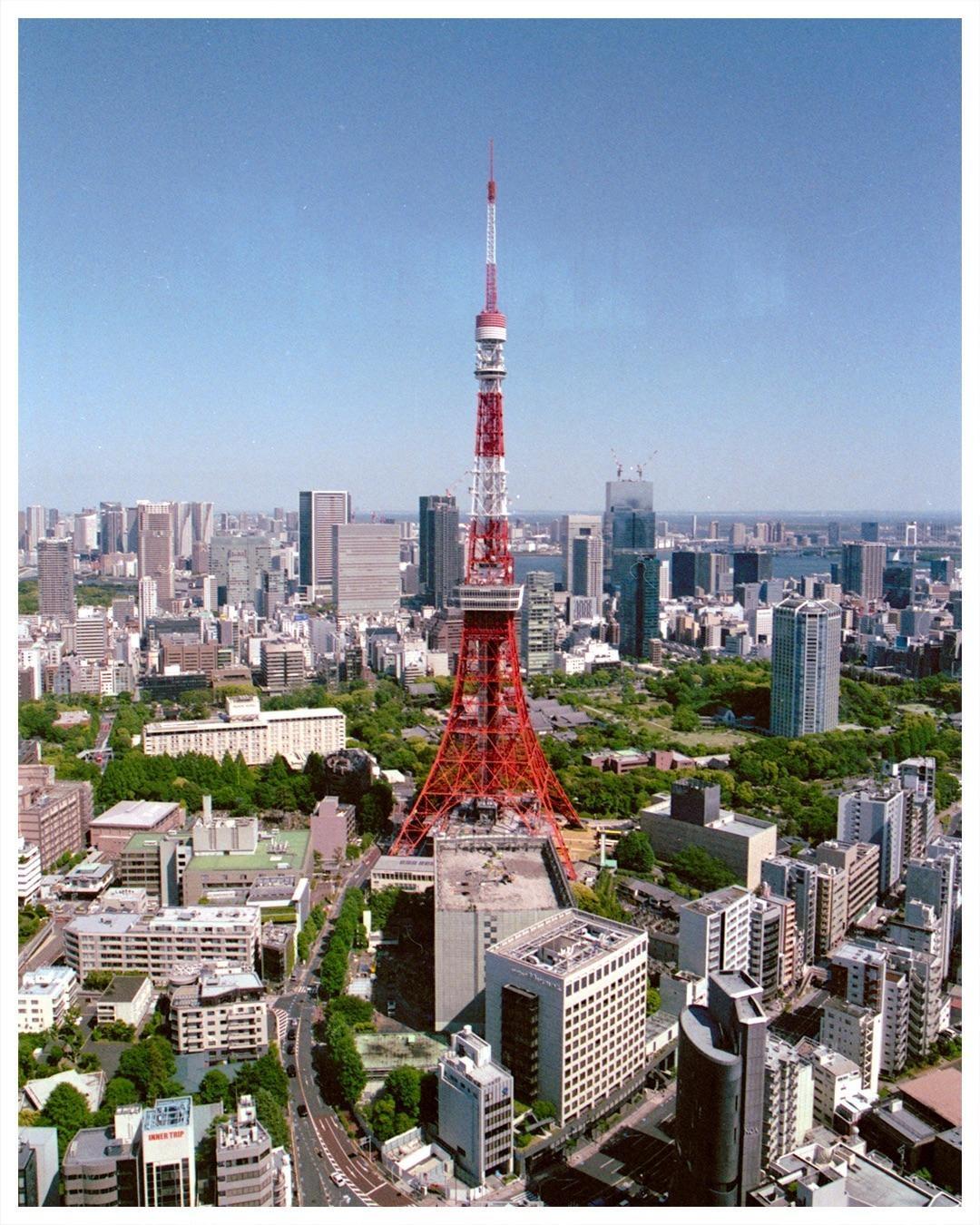 An aerial view shows the Tokyo Tower surrounded by a dense urban landscape with numerous buildings and green spaces under a clear sky.