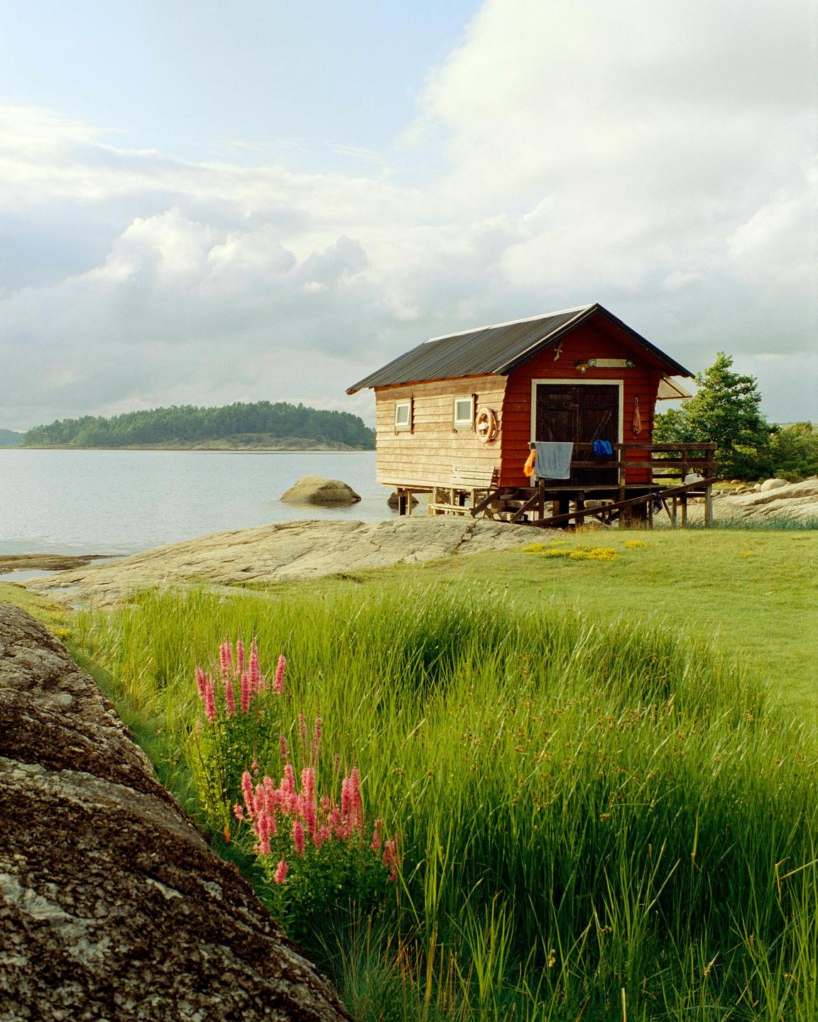 A red and wood cabin stands on stilts over a rocky shore next to a body of water with an island in the distance.