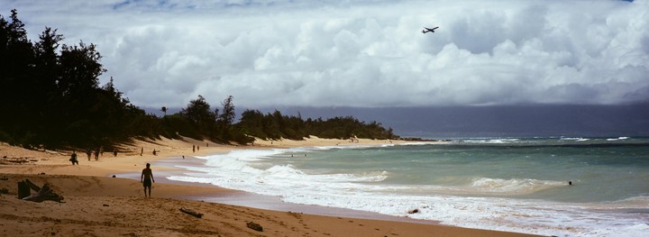 A small plane flies over a beach with people walking on the sand and swimming in the ocean under a cloudy sky.