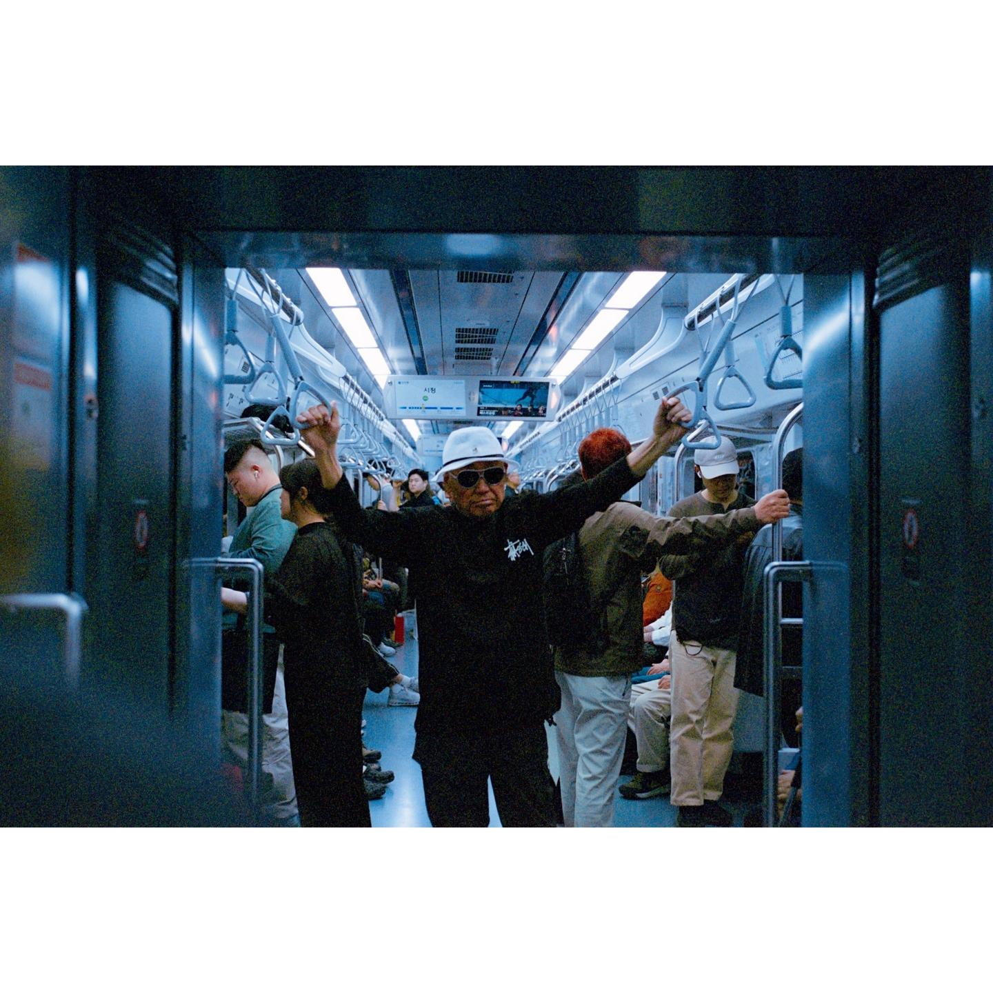 A man in sunglasses and a white hat stands with his arms raised in a subway car full of people.