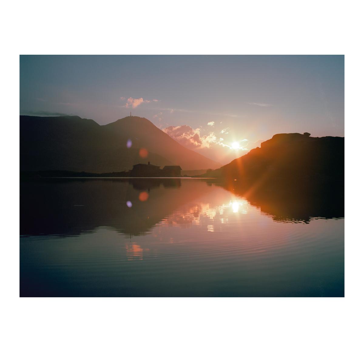 The sun sets behind mountains, casting a reflection on the water where a building is visible.