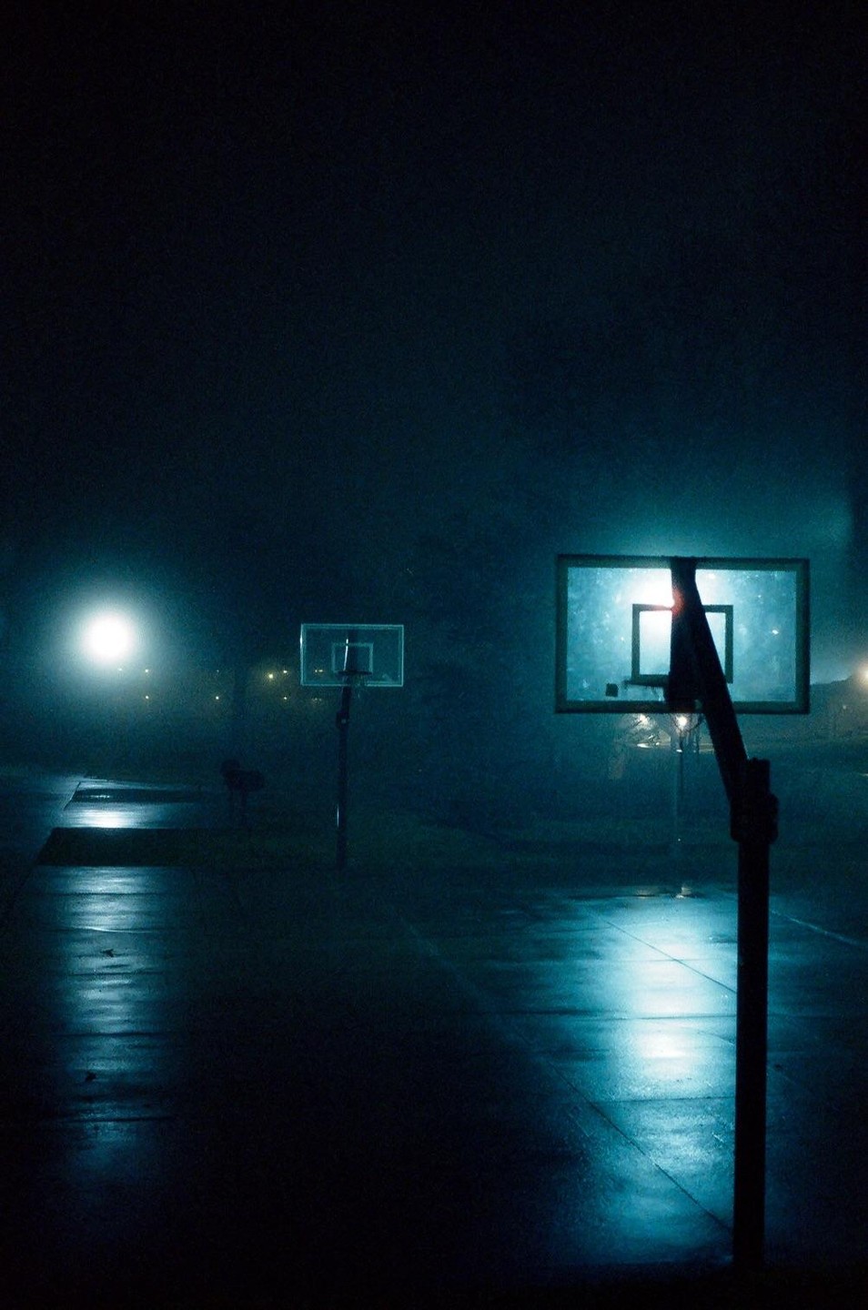 Two basketball hoops stand on a court at night, illuminated by a bright light source.