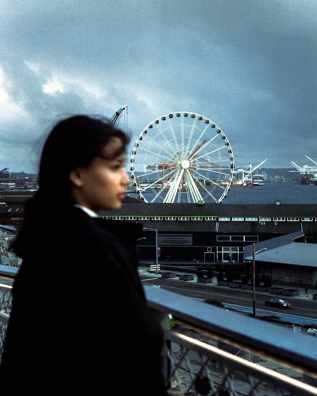 A woman in profile looks out over a city street and waterfront with a large Ferris wheel under a cloudy sky.