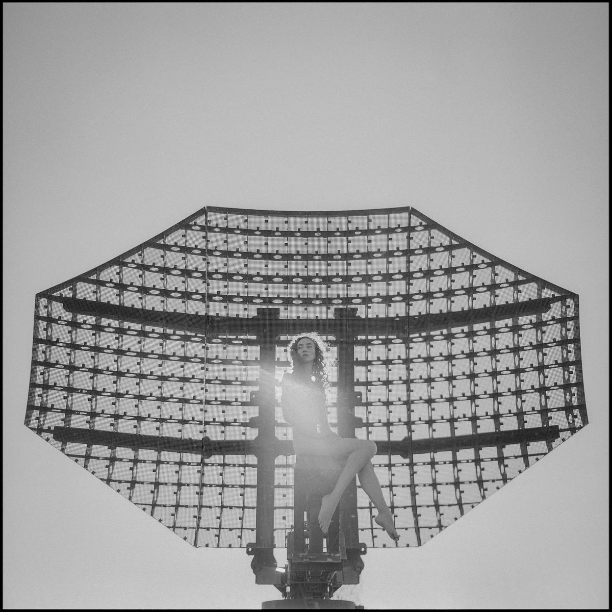 A woman sits on a large radar dish against a bright sky.