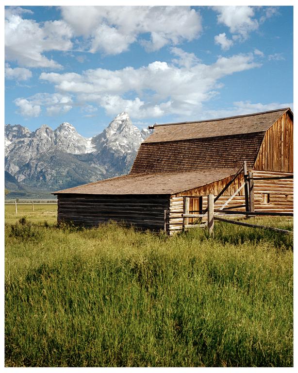 A wooden barn stands in a field of tall grass with mountains in the background under a cloudy sky.