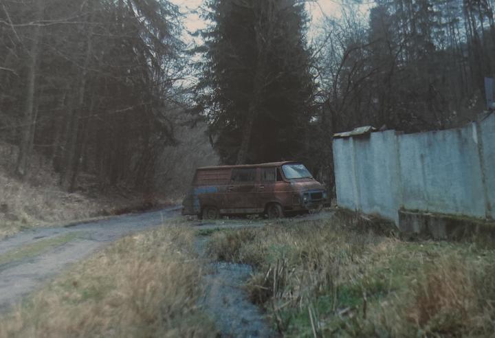 A rusty van is parked on a dirt road next to a concrete wall and a forest.