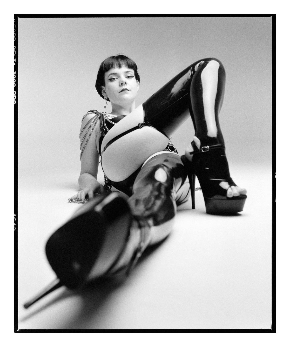 A woman in latex clothing and high heels is lying on the floor, looking at the viewer.