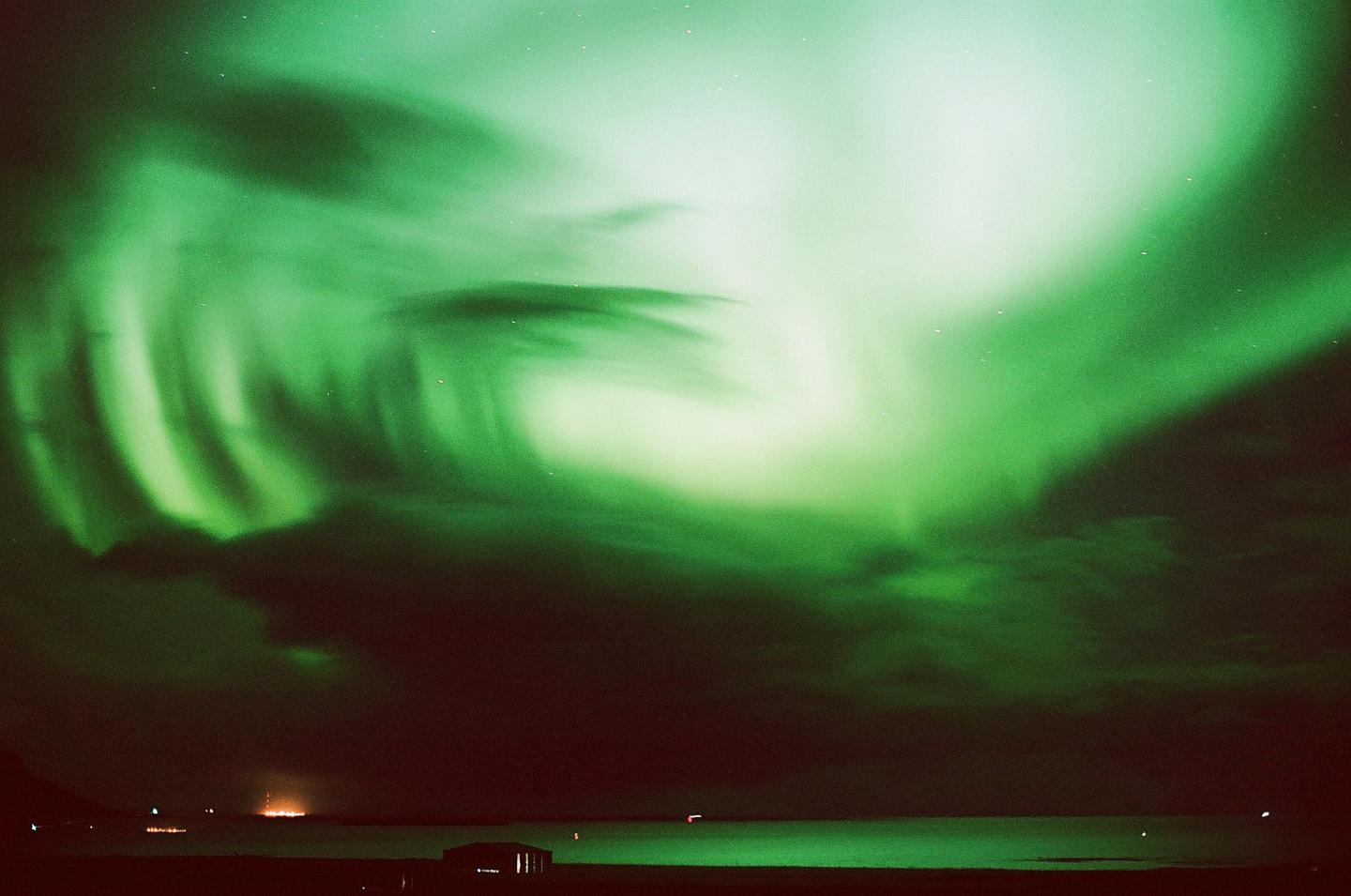 The northern lights glow green over a body of water with a distant light source on the horizon.