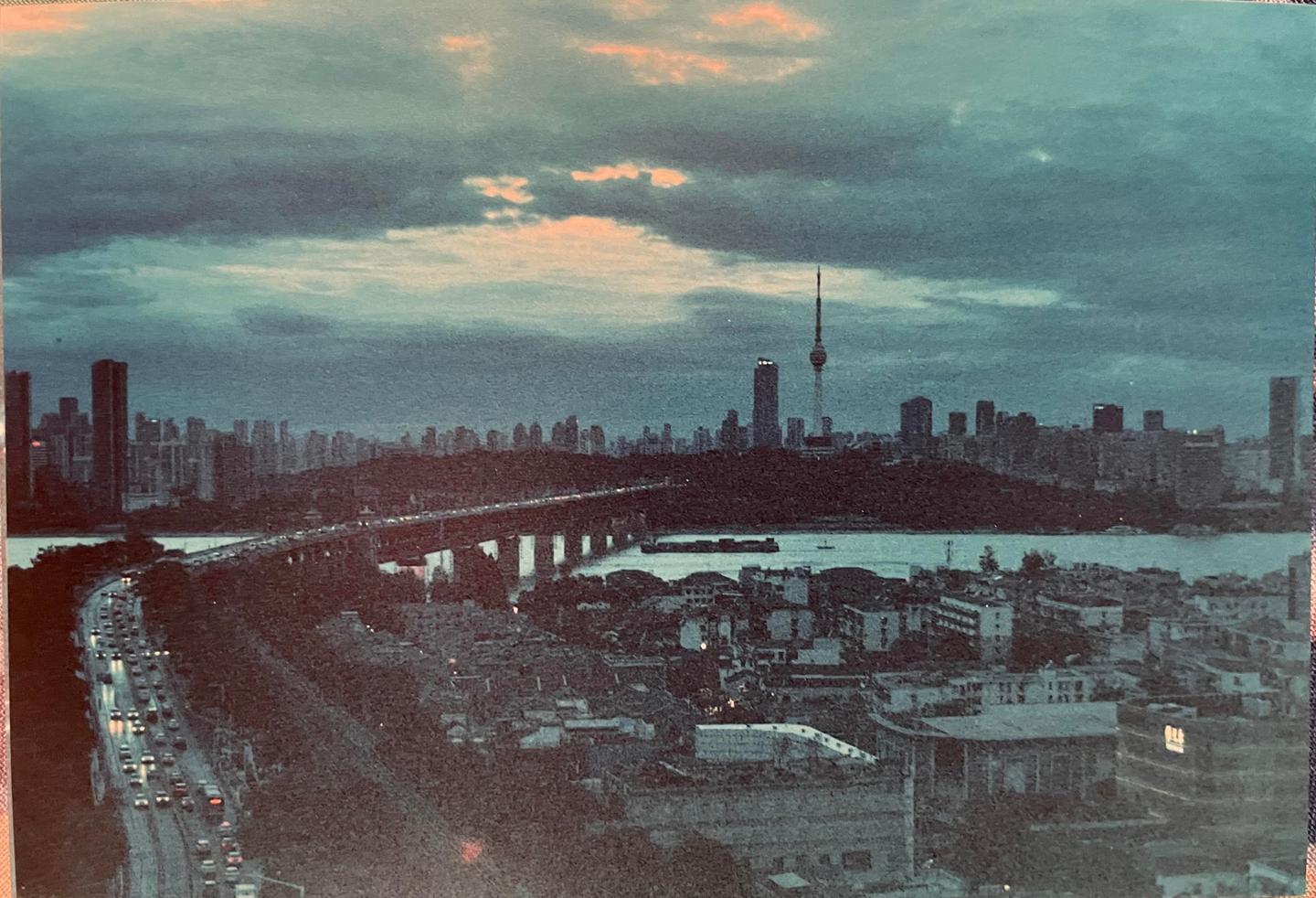 A city skyline with a river and a bridge is visible under a cloudy sky.