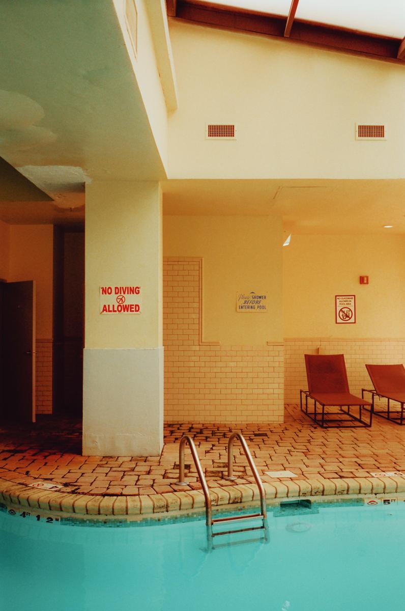 A swimming pool with a ladder and two lounge chairs is visible in an indoor setting with yellow walls and brick-patterned flooring.