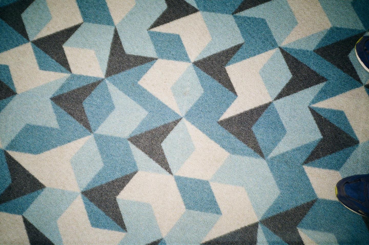 A blue shoe is visible on a patterned carpet with geometric shapes in shades of blue, gray, and white.