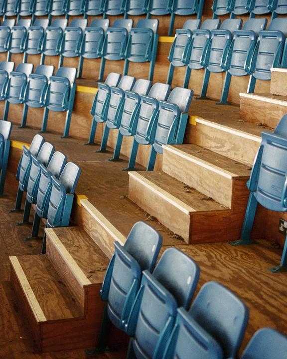 Rows of empty blue stadium seats are arranged on wooden risers.