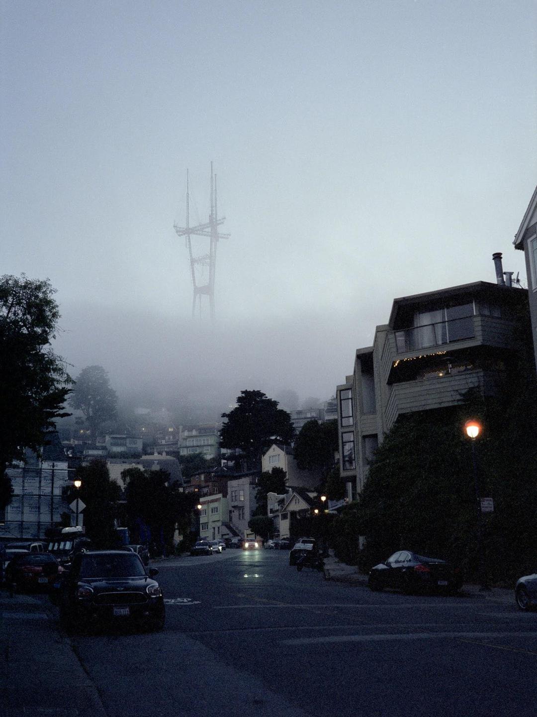 A street with parked cars and houses on a foggy evening, with the top of a tower visible through the fog.