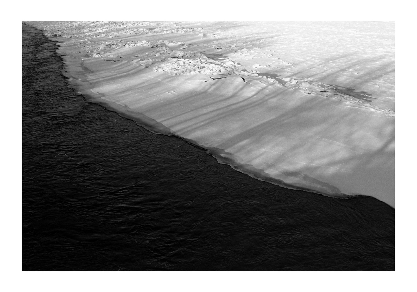 A river with dark water flows alongside a snow-covered bank with long shadows stretching across it.