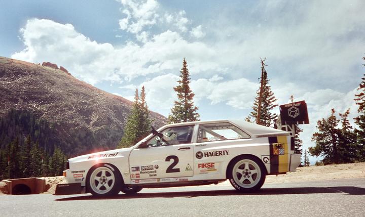 A white race car with the number two on its side is parked on a road with a mountain and pine trees in the background.
