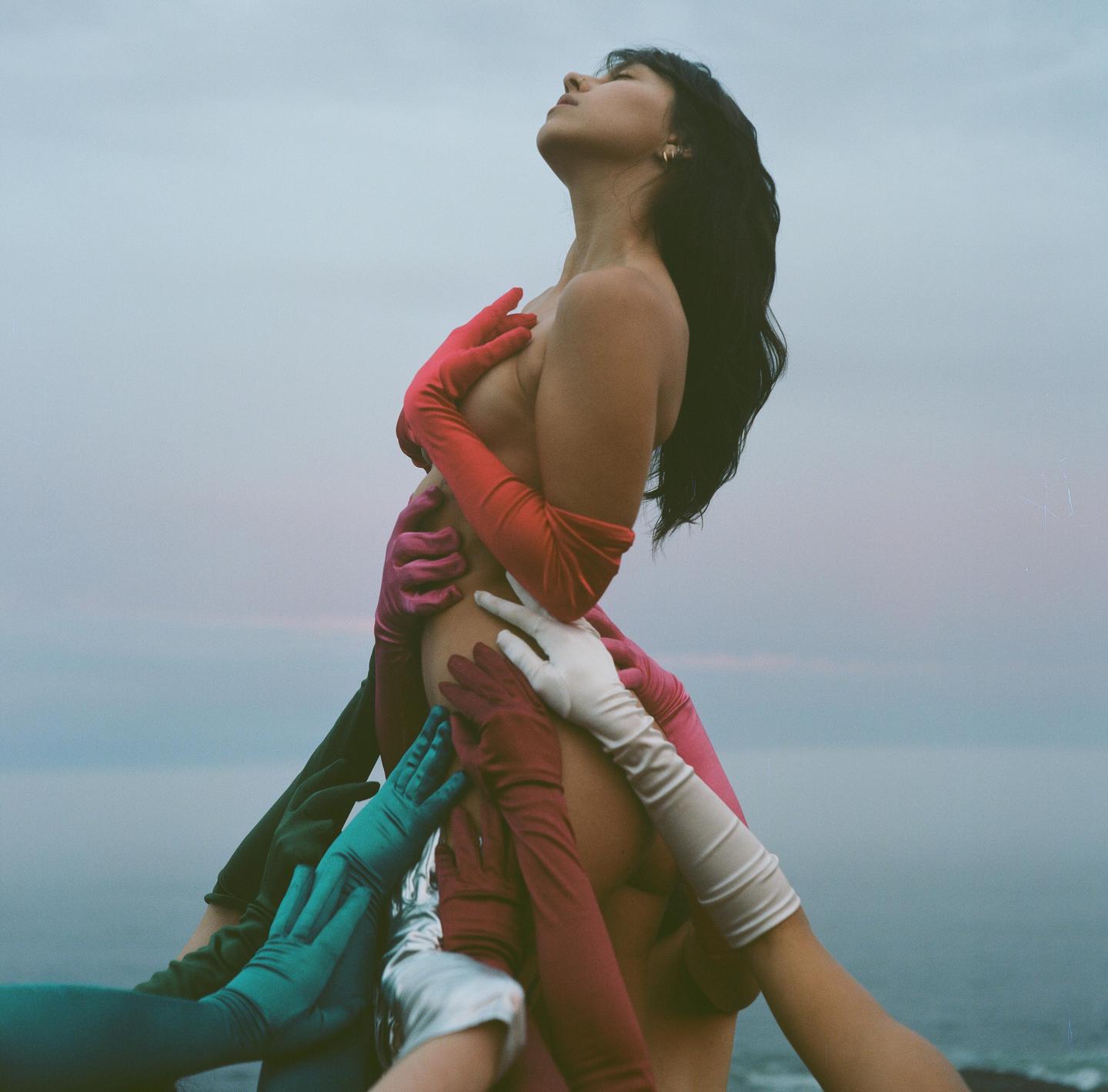 A woman with her eyes closed and head tilted back is surrounded by many gloved hands touching her body.