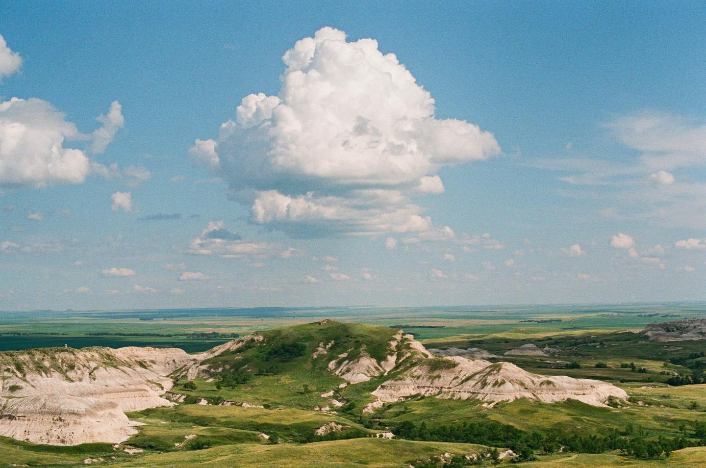 A vast landscape of rolling green hills and white rock formations stretches under a bright blue sky with fluffy white clouds.