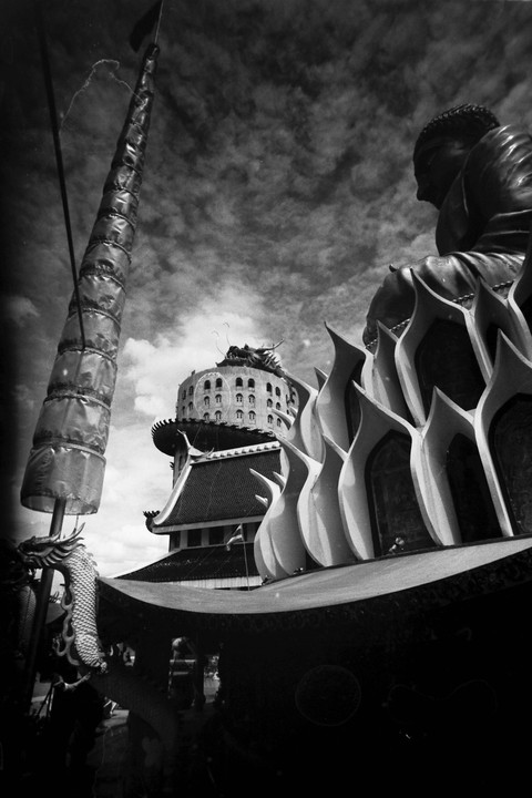 A low-angle, black and white shot shows a large Buddha statue and a tall, cylindrical structure with a dragon sculpture at its base, set against a cloudy sky.
