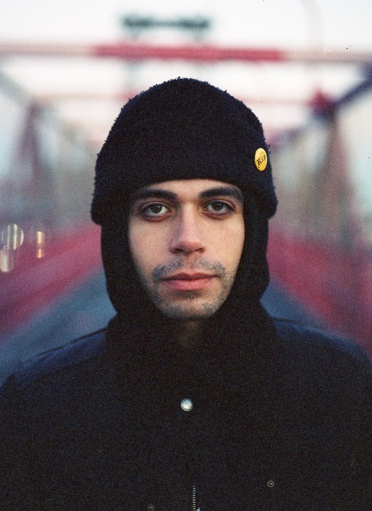 A man wearing a black beanie and jacket looks directly at the viewer.