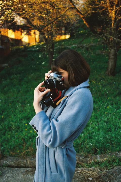 A woman in a blue coat holds a camera to her eye, standing in a grassy area with trees in the background.
