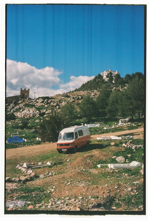 A white and orange van is parked on a dirt road in a hilly landscape with a building on a distant hilltop.