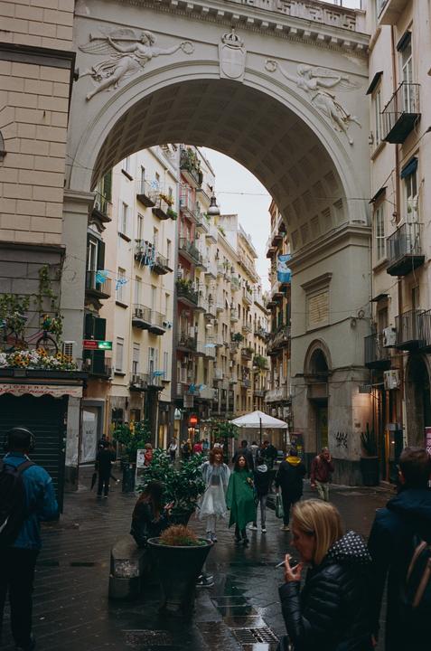 A crowd of people walk along a narrow street under a large archway in a city.