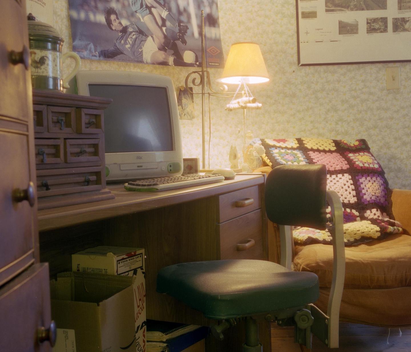 A desk with a computer, keyboard, and lamp is next to an armchair with a colorful blanket.