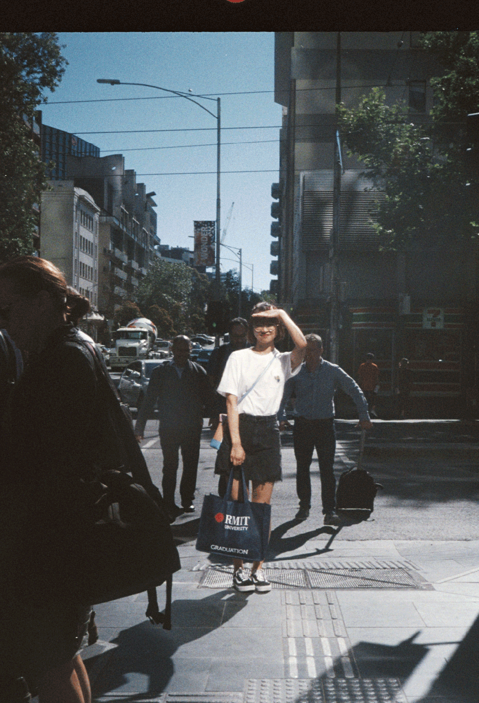 A woman shields her eyes from the sun while standing on a city street with other pedestrians and buildings in the background.
