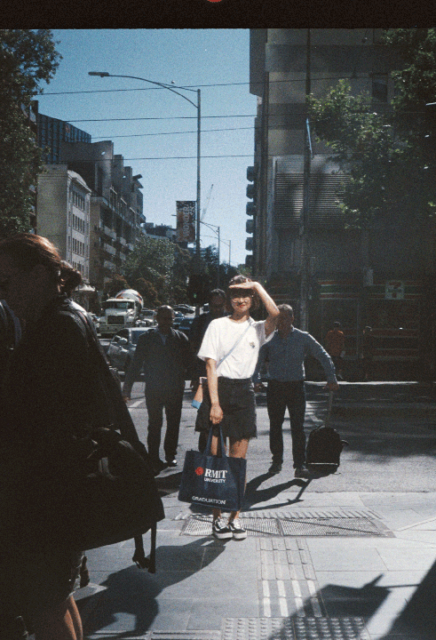A woman shields her eyes from the sun while standing on a city street with other pedestrians and buildings in the background.