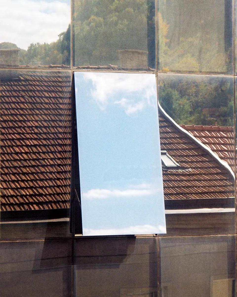 A mirror reflects the sky and clouds, surrounded by reflections of a tiled roof and trees.