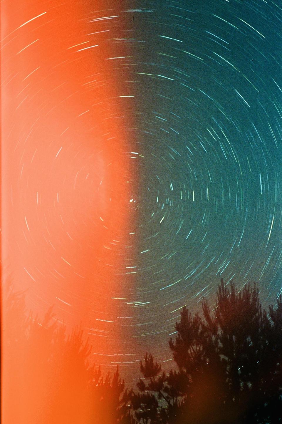 Star trails are visible in the night sky above trees, with a strong orange light source on the left.