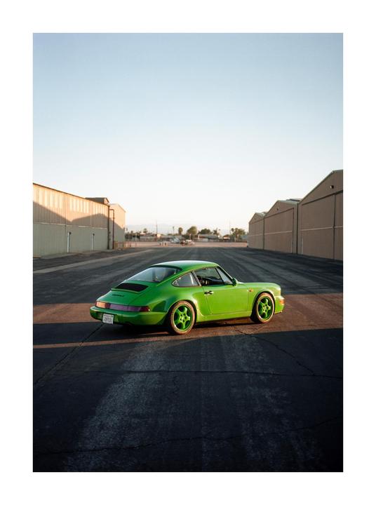 A bright green sports car is parked on an asphalt lot between two rows of hangars.