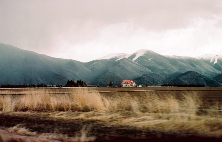 A white house with a red roof stands in a field with mountains in the background under a cloudy sky.