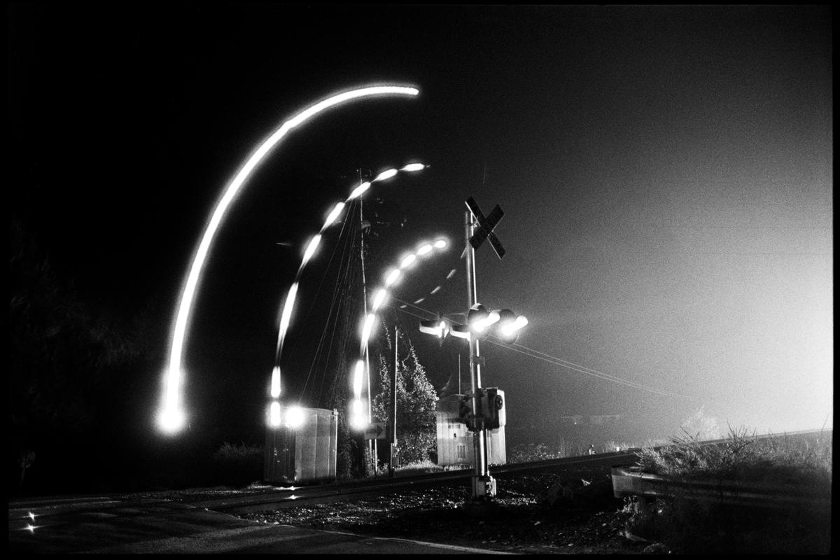 A black and white long exposure photograph captures light trails from a moving train at a railroad crossing at night.