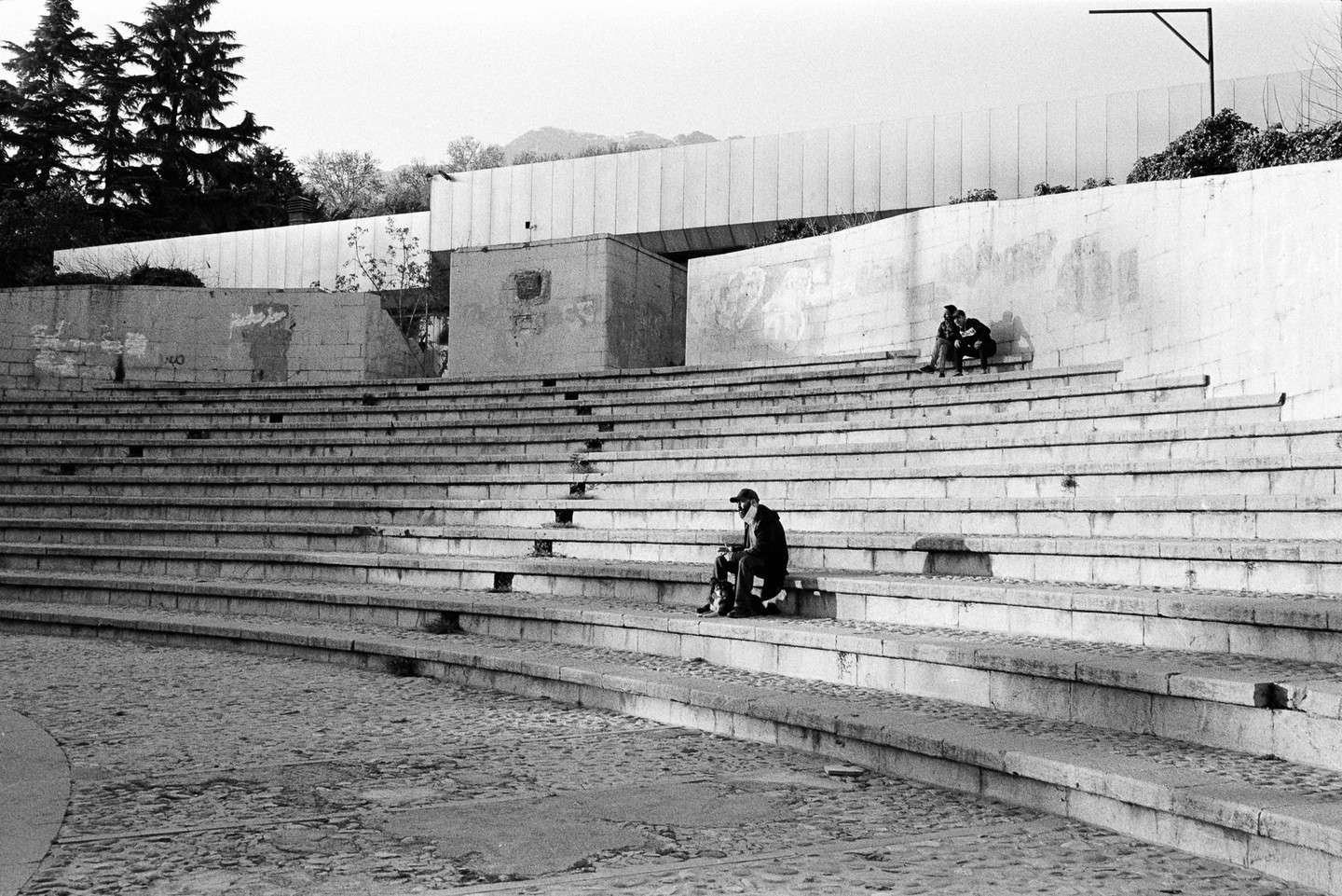 A man sits alone on the lower steps of an amphitheater while a couple sits together higher up.