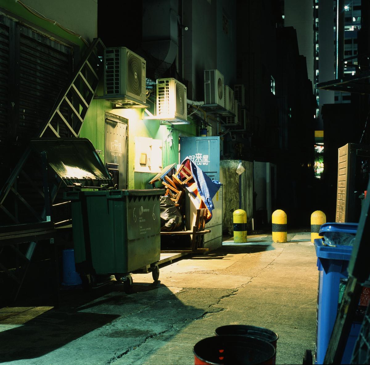 A green dumpster sits in a narrow alleyway at night, illuminated by a bright light.