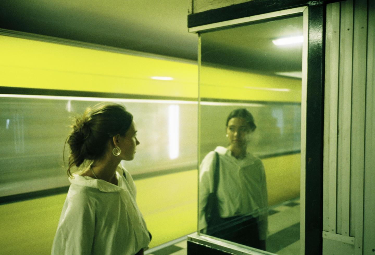 A woman with her hair tied back looks at her reflection in a glass panel.