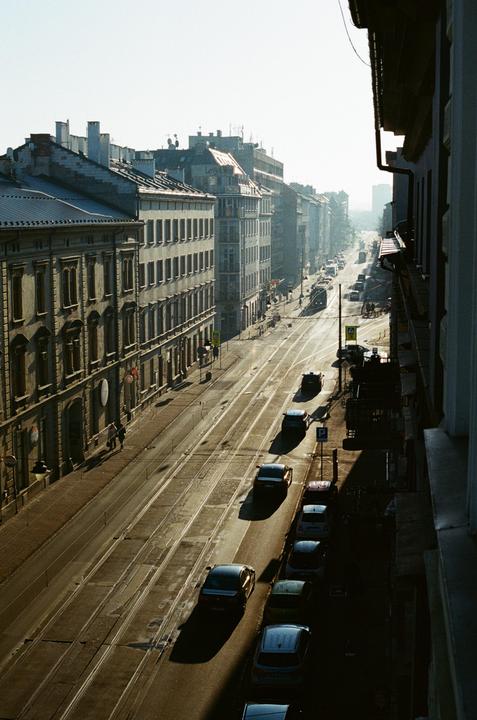 A long street with buildings on both sides and cars parked along the right side is illuminated by morning light.