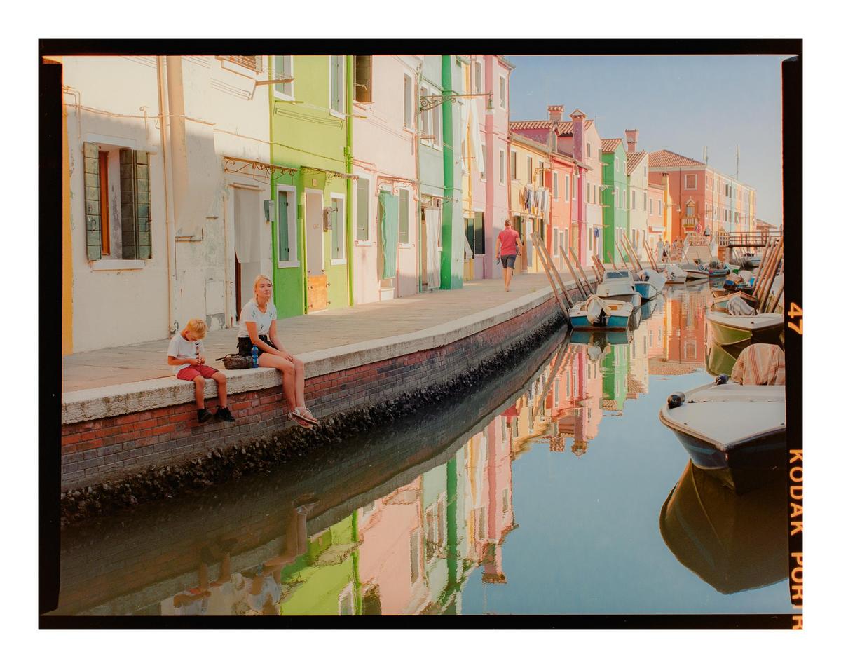 A woman and a child sit on the edge of a canal lined with colorful buildings and boats in Burano, Italy.