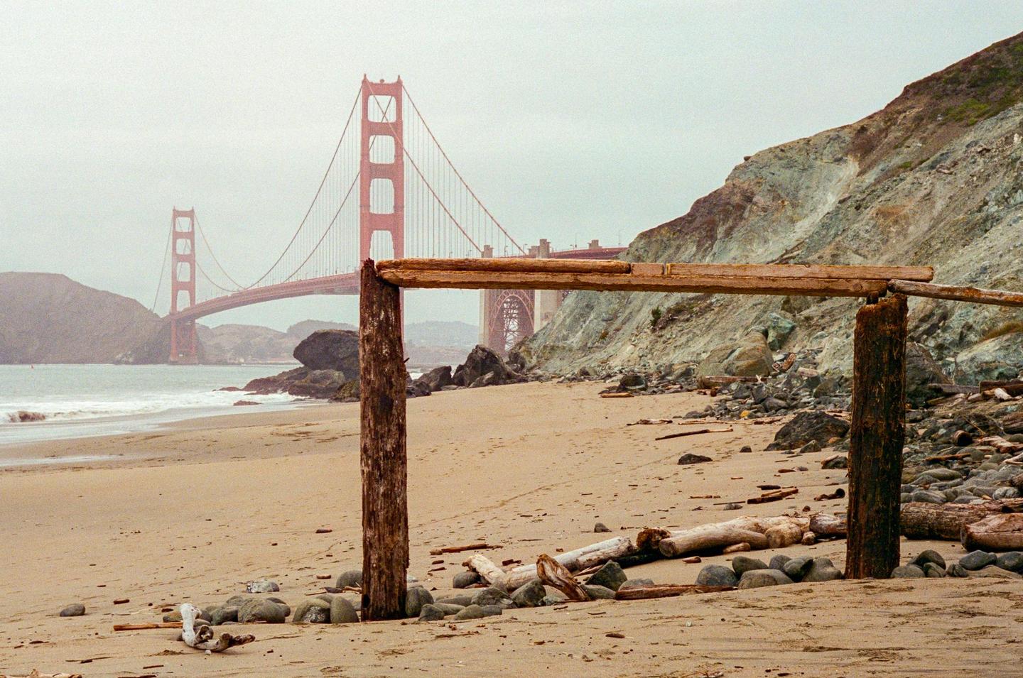 A wooden structure stands on a sandy beach with the Golden Gate Bridge visible in the distance under a hazy sky.