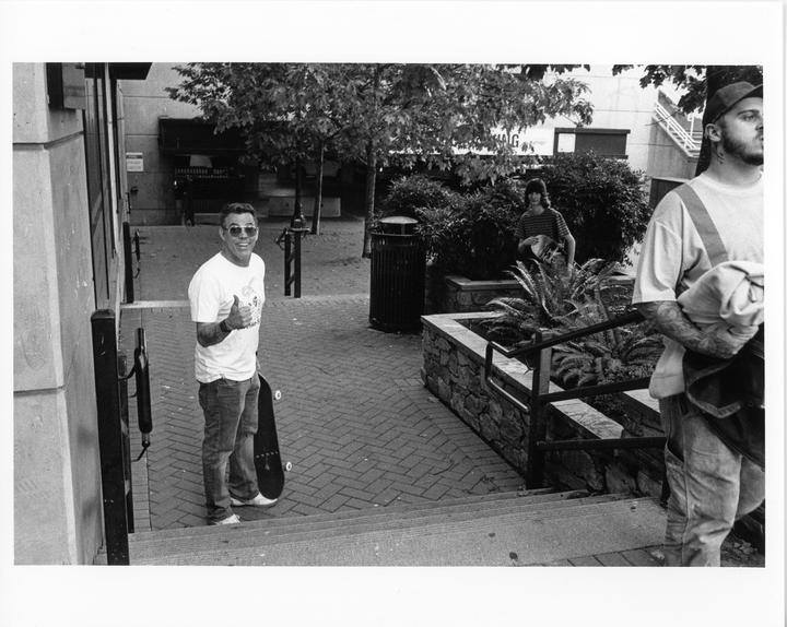 A man with a skateboard gives a thumbs up on a brick sidewalk next to a planter with lush foliage.