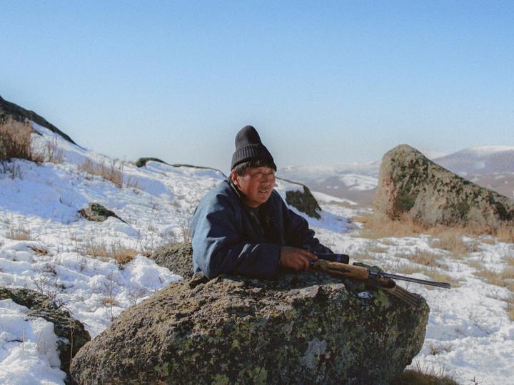 A man in a dark jacket and hat rests on a large rock with a rifle in a snowy, mountainous landscape.