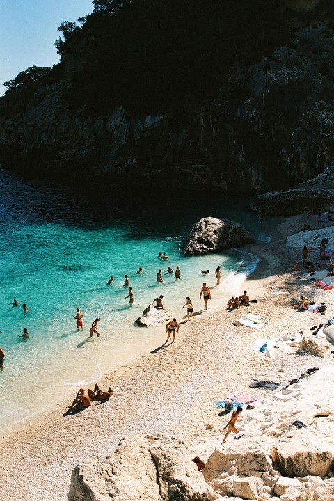 A group of people are swimming and relaxing on a sunny beach next to a large cliff.