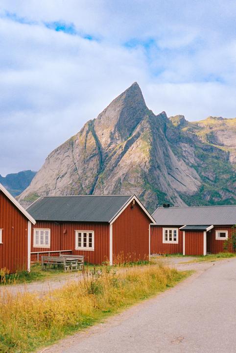 Red cabins with white trim line a dirt road with a large mountain in the background under a cloudy sky.